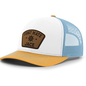 May include: A trucker hat with a white front panel, a light blue mesh back, and a yellow brim. The hat features a brown patch with the words "FIRST MATE JACE" and a ship's wheel design.
