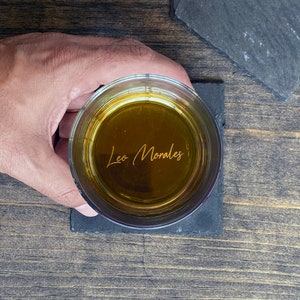 May include: A glass of amber liquid with the name "Leo Morales" engraved on the bottom. The glass is sitting on a black slate coaster.