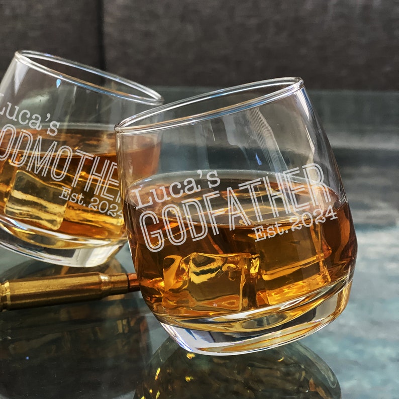May include: Two clear glass tumblers with the text "Luca's Godfather Est. 2024" etched on the side. The glasses are filled with amber liquid and ice cubes.