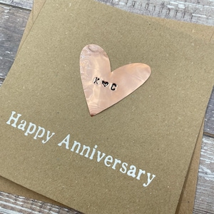 Puede incluir: Una tarjeta marr&oacute;n con el texto "Happy Anniversary" en letras blancas. Un coraz&oacute;n de cobre con las iniciales "K & G" est&aacute; unido a la tarjeta.