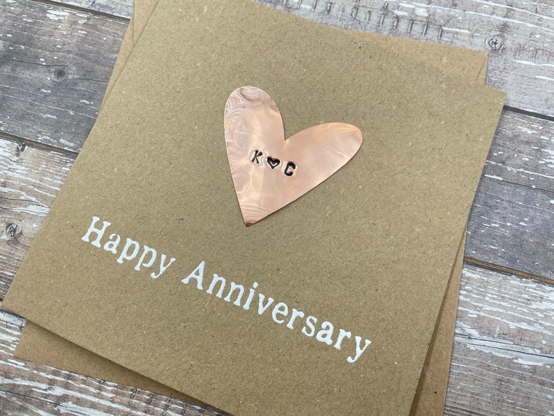 Puede incluir: Una tarjeta marr&oacute;n con el texto "Happy Anniversary" en letras blancas. Un coraz&oacute;n de cobre con las iniciales "K & G" est&aacute; unido a la tarjeta.