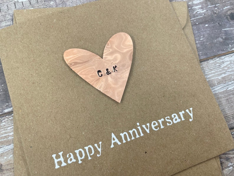 Puede incluir: Una tarjeta marr&oacute;n con un coraz&oacute;n de madera pegado. El coraz&oacute;n tiene las iniciales "C & K" talladas en &eacute;l. La tarjeta dice "Feliz Aniversario" en letras blancas.