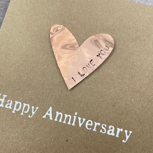 Puede incluir: Una tarjeta marr&oacute;n con el texto "Happy Anniversary" impreso en blanco. Un coraz&oacute;n de madera con el texto "I Love You" est&aacute; pegado a la tarjeta.