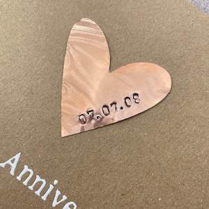 Puede incluir: Una tarjeta marr&oacute;n con las palabras "Happy Anniversary" impresas en blanco. Un coraz&oacute;n de cobre con la fecha "07.07.08" grabada en &eacute;l est&aacute; unido a la tarjeta.
