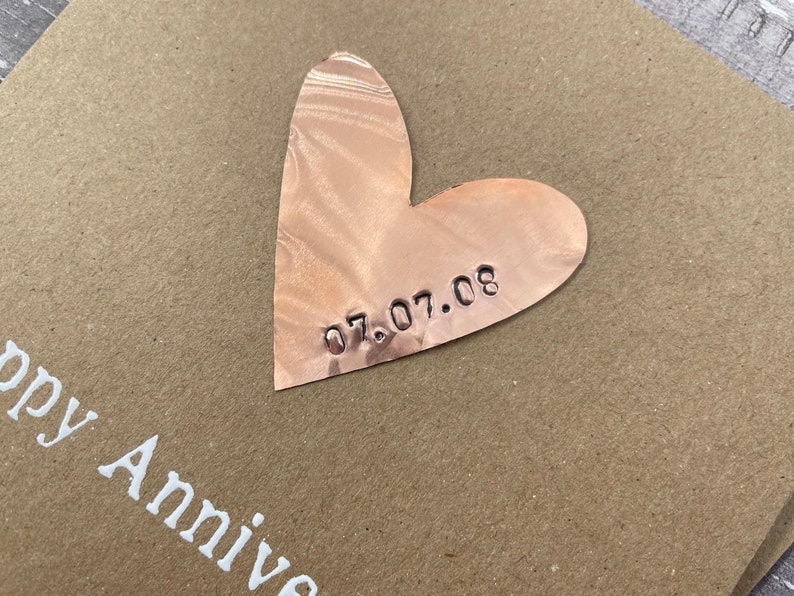 Puede incluir: Una tarjeta marr&oacute;n con las palabras "Happy Anniversary" impresas en blanco. Un coraz&oacute;n de cobre con la fecha "07.07.08" grabada en &eacute;l est&aacute; unido a la tarjeta.