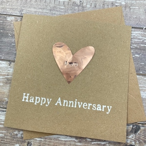 Puede incluir: Una tarjeta marr&oacute;n con el texto "Happy Anniversary" en letras blancas. Un coraz&oacute;n de cobre con el texto "I &hearts; U" est&aacute; unido a la tarjeta.
