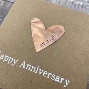 Puede incluir: Una tarjeta marr&oacute;n con las palabras "Happy Anniversary" impresas en blanco. Un coraz&oacute;n de cobre con la fecha "07.07.08" grabada en &eacute;l est&aacute; unido a la tarjeta.
