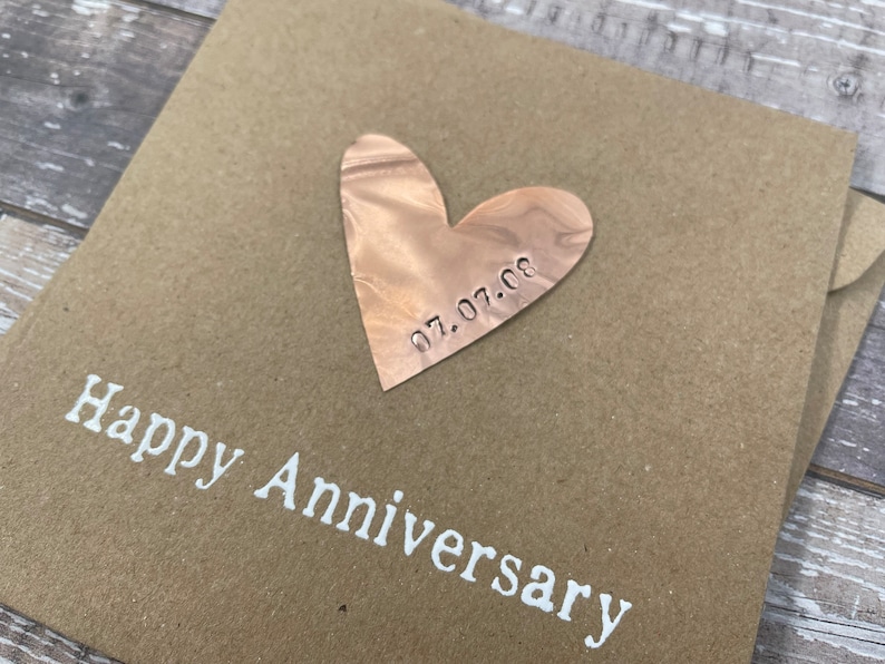 Puede incluir: Una tarjeta marr&oacute;n con las palabras "Happy Anniversary" impresas en blanco. Un coraz&oacute;n de cobre con la fecha "07.07.08" grabada en &eacute;l est&aacute; unido a la tarjeta.