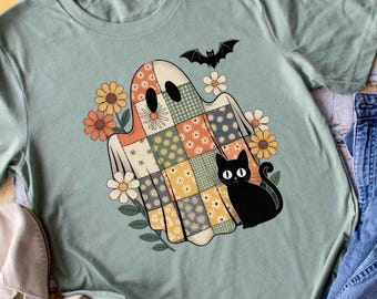 May include: A light green t-shirt featuring a patchwork ghost design with a black cat, flowers, and a bat. The shirt is paired with blue jeans, a wooden bead necklace, and beige shoes, creating a casual, fall-themed ensemble.