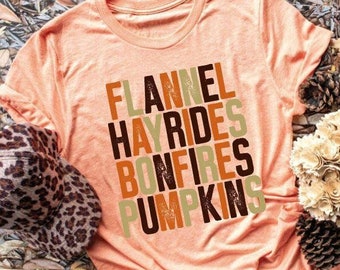 May include: Peach-colored t-shirt with the words "FLANNEL HAYRIDES BONFIRES PUMPKINS" in a stacked design. A leopard print hat and a small pumpkin are next to the shirt. The background includes leaves and pinecones.