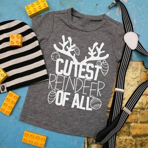 May include: A gray t-shirt with the text "Cutest Reindeer of All" in white, featuring a reindeer with antlers and Christmas ornaments. The shirt is paired with a black and white striped beanie and black and white striped suspenders.