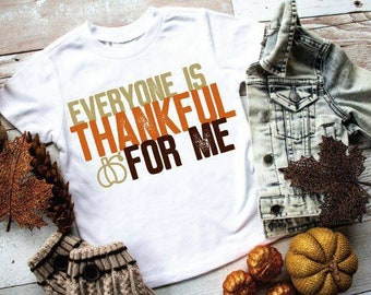 May include: White t-shirt with the text "EVERYONE IS THANKFUL FOR ME" in brown, orange, and tan. A denim jacket, decorative pumpkins, and fall foliage are arranged around the shirt on a wooden surface.