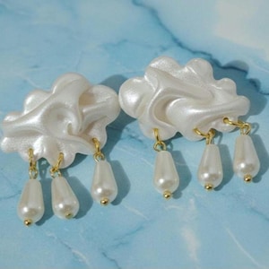 Può includere: Un paio di orecchini a forma di nuvola con finitura perlata. Ogni orecchino presenta tre finte perle a goccia sospese da ganci color oro. Sfondo blu e bianco marmorizzato.