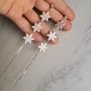 Könnte beinhalten: Ein Paar silberne Schneeflockenohrringe mit weißen Perlenakzenten. Die Ohrringe sind an einer Kette und hängen am Ohrläppchen.