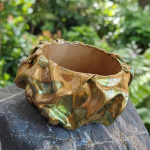 Pode incluir: Uma pulseira de punho artesanal com a parte superior aberta. O exterior é um dourado metálico texturizado com detalhes verdes. O interior é castanho claro. A pulseira está sobre uma pedra cinzenta, com um fundo verde desfocado.