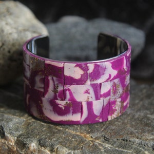 Könnte beinhalten: Ein breites Armband mit Mosaikdesign. Das Armband hat eine silberne Innenseite und ist mit einem Muster aus rosa, weißen und goldfarbenen rechteckigen Fliesen verziert. Das Armband wird auf einer strukturierten Steinoberfläche präsentiert.