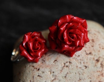 May include: Two red rose-shaped rings made of polymer clay. The rings are on a light brown textured surface.