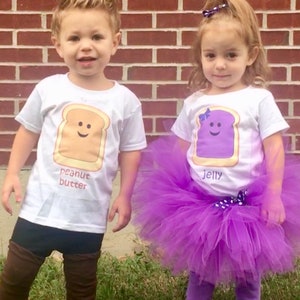 May include: Two children dressed in matching white t-shirts with food-themed graphics. One shirt says "peanut butter" with a smiling peanut butter sandwich. The other says "Jelly" with a smiling jelly sandwich and a purple tutu.