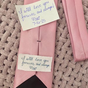 May include: A light pink necktie with a black lining. A handwritten note is attached, reading "I will love you forever and always!" with a name and date. The tie is lying on a textured, light purple surface.