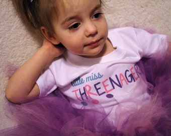 May include: A young girl wearing a white shirt with the text "little miss three-nager" and a purple tulle skirt. She has a purple glitter bow in her hair.