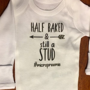 May include: White long-sleeve baby onesie with black text that reads "Half Baked & still a stud #micropreemie".