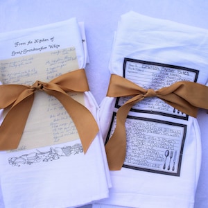 May include: Two white tea towels with brown ribbon tied around them. One towel has a handwritten recipe for "Mango Cake" and the text "From the Kitchen of Great Grandmother White". The other towel has a printed recipe for "Chicken Cheese Bites".