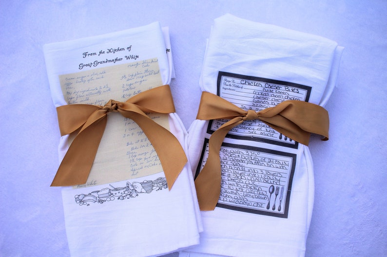 May include: Two white tea towels with brown ribbon tied around them. One towel has a handwritten recipe for "Mango Cake" and the text "From the Kitchen of Great Grandmother White". The other towel has a printed recipe for "Chicken Cheese Bites".