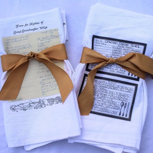 May include: Two white kitchen towels with handwritten recipes printed on them. One towel has a recipe for "Chicken Caesar Bites" and the other has a recipe from "Great Grandmother Wije". Both towels are tied with a brown ribbon.