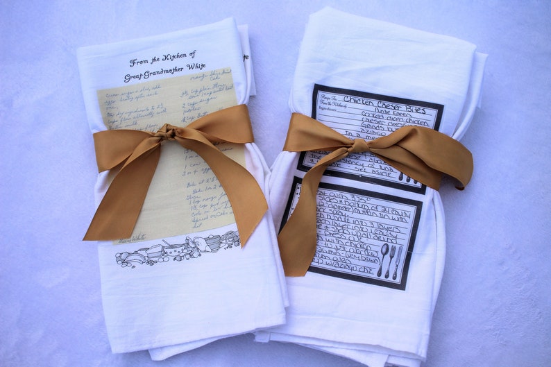 May include: Two white kitchen towels with handwritten recipes printed on them. One towel has a recipe for "Chicken Caesar Bites" and the other has a recipe from "Great Grandmother Wije". Both towels are tied with a brown ribbon.