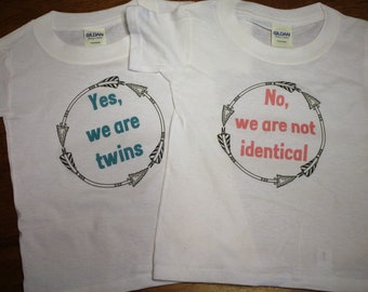 May include: Two white t-shirts with a black arrow design around the text. One shirt says "Yes, we are twins" in teal text. The other shirt says "No, we are not identical" in pink text.