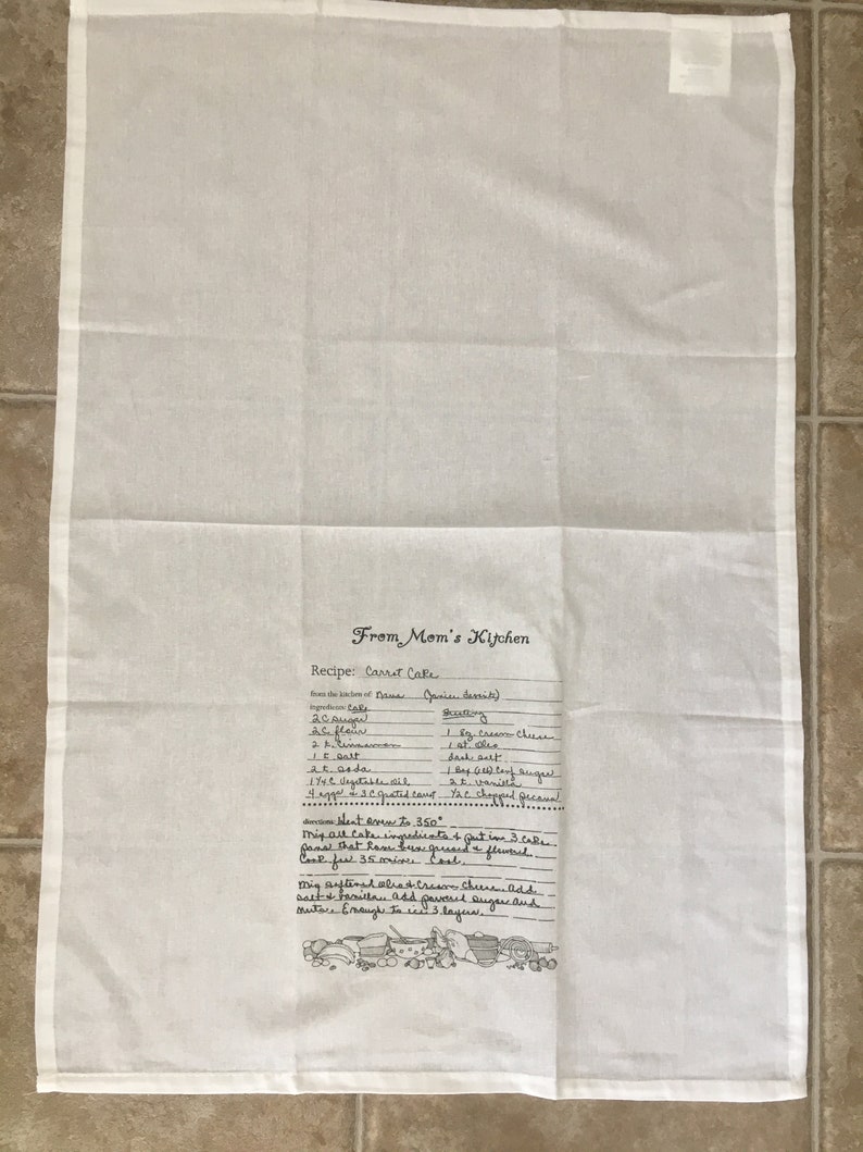 May include: A white kitchen towel with a handwritten recipe for carrot cake. The recipe includes ingredients like carrots, sugar, flour, and spices. The towel also features a small illustration of kitchen tools.