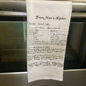 May include: A white kitchen towel with a handwritten recipe for carrot cake from "From Mom's Kitchen". The recipe includes ingredients for the cake and frosting, as well as directions for baking.