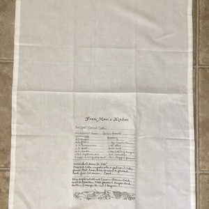 May include: A white kitchen towel with a handwritten recipe for carrot cake. The recipe includes ingredients like carrots, sugar, flour, and spices. The towel also features a small illustration of kitchen tools.