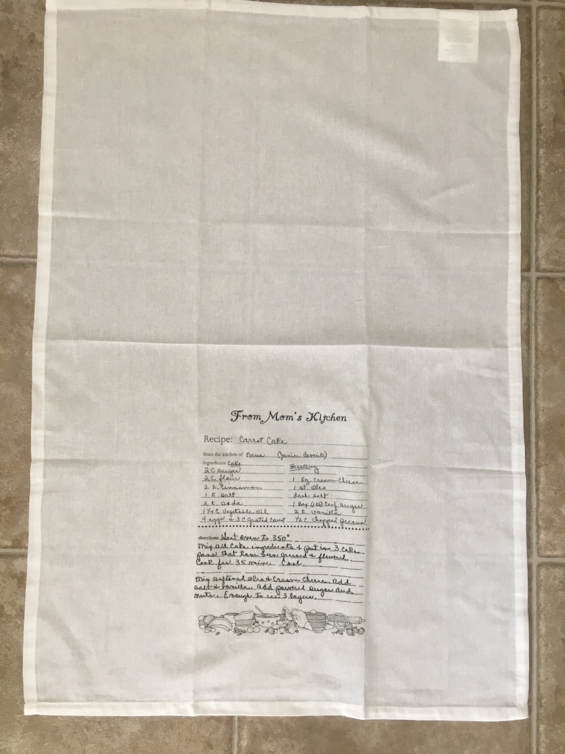 May include: A white kitchen towel with a handwritten recipe for carrot cake. The recipe includes ingredients like carrots, sugar, flour, and spices. The towel also features a small illustration of kitchen tools.