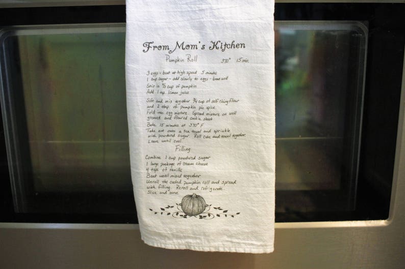 May include: A white tea towel with handwritten instructions for making a pumpkin roll. The instructions include ingredients and steps for both the pumpkin roll and the filling. The text reads "From Mom's Kitchen Pumpkin Roll 350&deg; 15 mins".