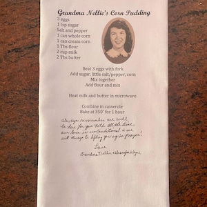 May include: A white kitchen towel with a handwritten recipe for Grandma Nellie's Corn Pudding. The recipe includes ingredients like corn, eggs, milk, and butter. The towel also has a photo of Grandma Nellie and a handwritten message at the bottom.