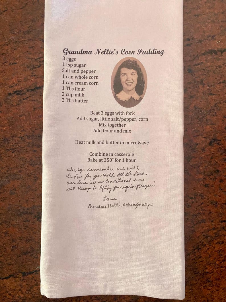 May include: A white kitchen towel with a handwritten recipe for Grandma Nellie's Corn Pudding. The recipe includes ingredients like corn, eggs, milk, and butter. The towel also has a photo of Grandma Nellie and a handwritten message at the bottom.
