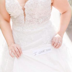 May include: A white wedding dress with lace detailing. The bride is holding up the skirt to reveal a handwritten note that says "I love you".
