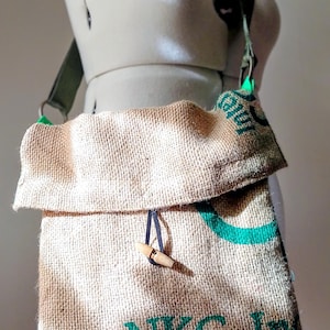 Puede incluir: Un bolso de hombro hecho de material de saco de café de arpillera color canela con texto impreso en verde que dice NKG Inc. El bolso incluye una solapa plegable asegurada por un cordón oscuro y un cierre de palanca de madera clara.