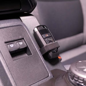 May include: A black car key fob secured by a black strap on a gray dashboard. The fob has several buttons and a small screen. Two window control buttons are visible on the dashboard.