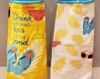 May include: Two crocheted kitchen towel holders with blue and white crocheted tops. The towel on the left has a yellow background with the text "Drink in hand toes in the sand" and a cocktail glass and flip flops. The towel on the right has a white background with yellow palm leaves and flip flops.