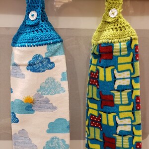 May include: Two crocheted tea towels hanging on a white oven door. The towel on the left has a blue crocheted top and a white background with a blue cloud pattern. The towel on the right has a green crocheted top and a blue background with a yellow and red boot pattern.