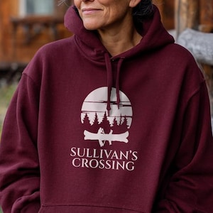 May include: A maroon hoodie with a white graphic featuring a mountain scene and the text "Sullivan's Crossing." The hoodie has a drawstring hood and a front pocket. The person is standing outside a wooden cabin.
