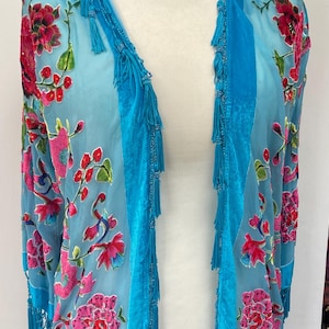 Op de afbeelding: Een doorschijnende, turquoise kimono-achtige kledingstuk met een bloemenpatroon in roze en rood. Het kledingstuk heeft turquoise fluwelen panelen en is versierd met turquoise kwastjes en kralen, wat een elegant, vloeiend ontwerp creëert.
