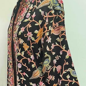 Könnte beinhalten: Eine schwarze bestickte Jacke mit einem floralen und Vogel-Design. Die Jacke hat einen Stehkragen und lange Ärmel. Die Stickerei ist detailliert und kunstvoll und zeigt Pfauen, Vögel und Blumen in Rosatönen, Orange, Grün und Blau.
