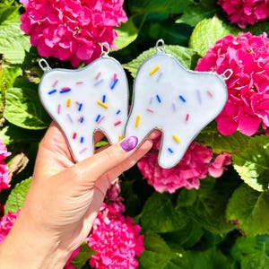 May include: Two white, tooth-shaped glass ornaments with colorful sprinkle designs. Each ornament has a silver-colored metal border and a small loop for hanging. The background features vibrant pink flowers and green foliage.