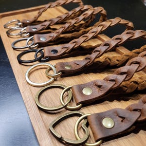 May include: A set of five leather keychains with braided straps and different metal key rings. The keychains are brown and have a rustic look.