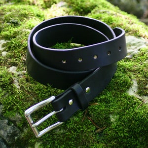 May include: A black leather belt with a silver buckle. The belt is folded in half and lying on a bed of green moss.