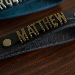 May include: A black leather strap with the name "MATTHEW" in gold metal letters. The strap has two brass rivets.