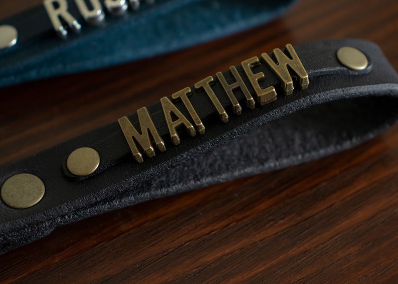 May include: A black leather strap with the name "MATTHEW" in gold metal letters. The strap has two brass rivets.